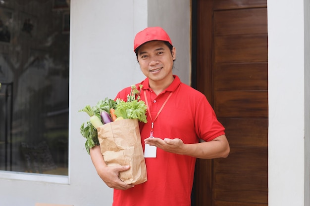 Livreur asiatique transportant une boîte de colis de légumes frais devant la porte de la maison