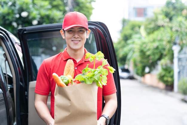 Livreur asiatique vêtu d'un uniforme rouge et chapeau rouge tenant un sac en papier de nourriture, de fruits, de légumes souriant et debout près de la porte de la voiture devant la maison.