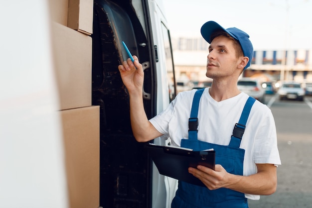 Livreur en cases à cocher uniformes dans la voiture, service de livraison. Homme, debout, à, emballages carton, dans, véhicule, homme, livrer