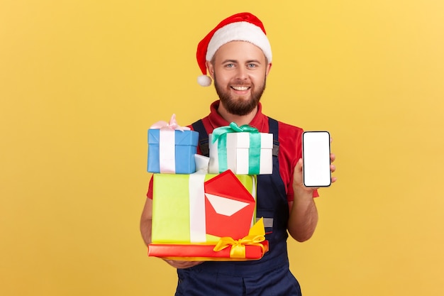 Livreur en chapeau de père noël rouge tenant des cadeaux de vacances et montrant un smartphone avec un affichage vide