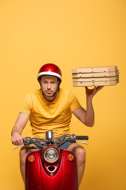 Livreur concentré en uniforme jaune faisant du scooter avec des boîtes à pizza isolées sur jaune