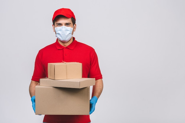 Livreur employé en casquette rouge t-shirt blanc uniforme masque facial gants tenir boîte en carton vide isolé sur mur blanc. Concept de virus de coronavirus pandémique de quarantaine de service 2019-ncov.