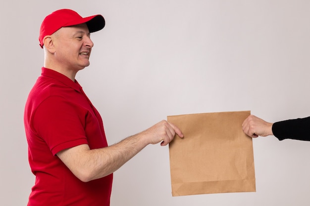 Livreur heureux et positif en uniforme rouge et casquette donnant un paquet de papier au client souriant amical