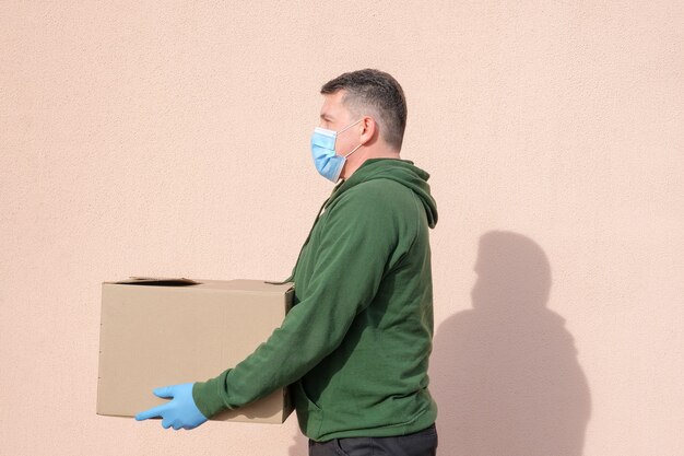 Livreur en masque médical et gants tenant et portant une boîte en carton