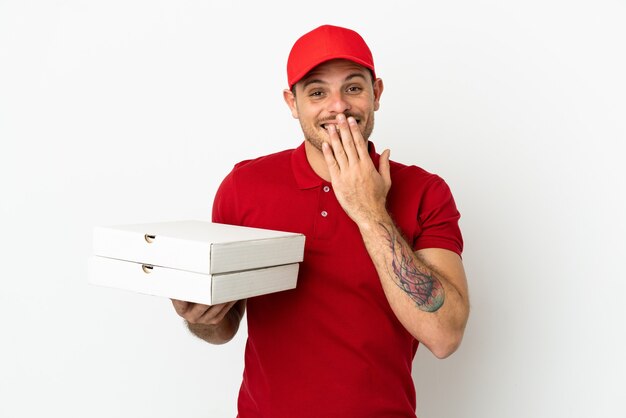 Livreur de pizza avec uniforme de travail ramassant des boîtes de pizza sur un mur blanc isolé heureux et souriant couvrant la bouche avec la main