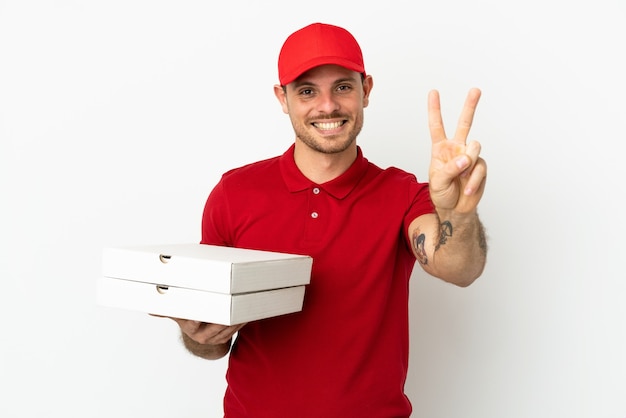 Livreur de pizza avec un uniforme de travail ramassant des boîtes de pizza sur un mur blanc isolé souriant et montrant le signe de la victoire