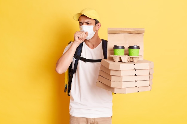 Livreur portant un masque de protection tenant des boîtes à pizza et un coursier de café à emporter travaillant pendant une épidémie de grippe attrapant un rhume ayant des symptômes de grippe debout isolé sur fond jaune