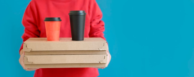 Photo livreur en t-shirt rouge, passant une commande de nourriture et tenant deux boîtes à pizza et des verres de café, sur fond bleu