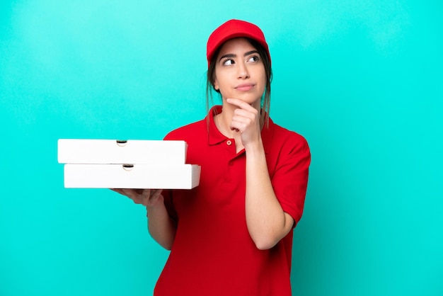 Livreuse de pizza avec uniforme de travail ramassant des boîtes de pizza isolées sur fond bleu ayant des doutes en levant les yeux