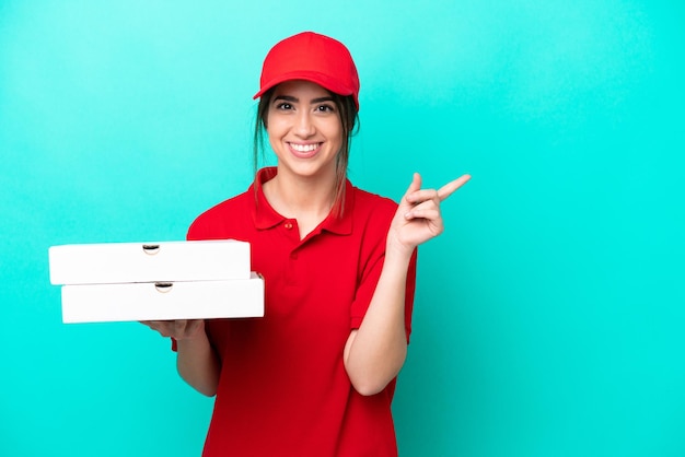 Livreuse de pizza avec uniforme de travail ramassant des boîtes de pizza isolées sur fond bleu pointant vers le côté pour présenter un produit