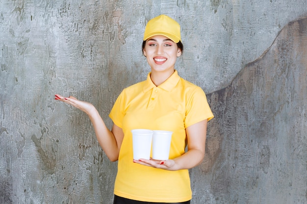 livreuse en uniforme jaune tenant deux gobelets en plastique de boisson et montrant quelque chose.