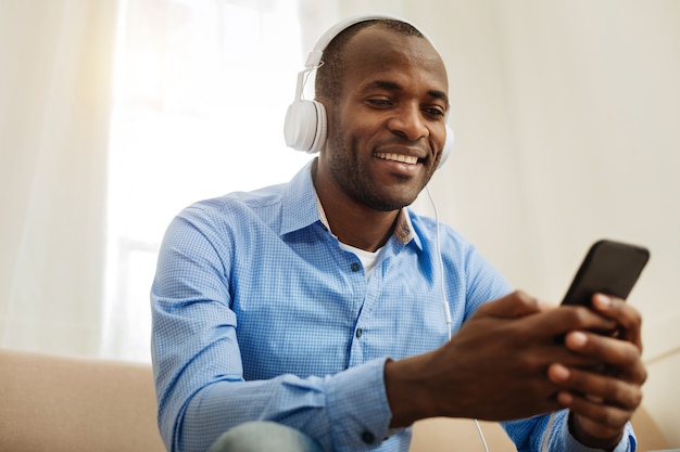 Loisir. Séduisant jeune homme aux cheveux noirs joyeux portant des écouteurs et tenant un téléphone et écouter de la musique et se détendre