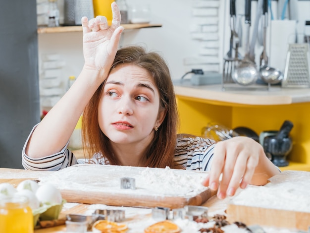 Loisirs culinaires. Portrait de dame s'ennuie assis dans la cuisine à table recouverte de farine, en attente de biscuits prêts.