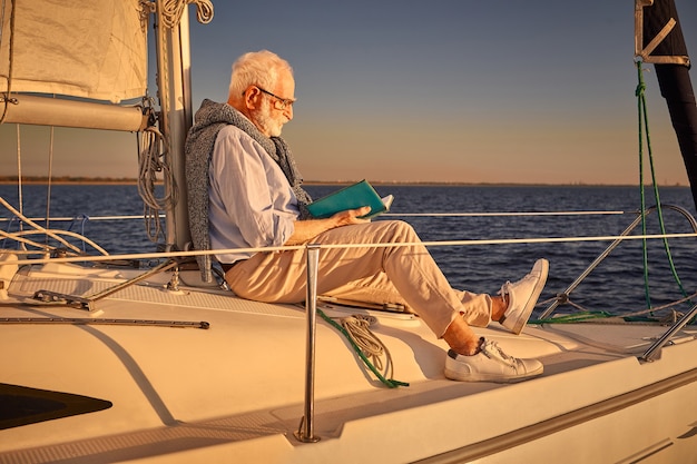 Loisirs et détente homme âgé lisant un livre assis sur le côté du voilier ou du pont du yacht
