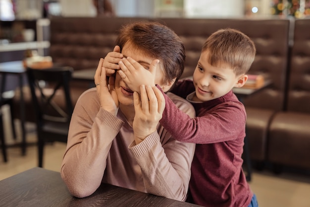 Loisirs et divertissements en famille. grand-mère heureuse avec des cheveux courts, des lunettes et un petit-fils se reposent dans un café. enfant embrasse grand-mère et ferme les yeux de ses propres mains