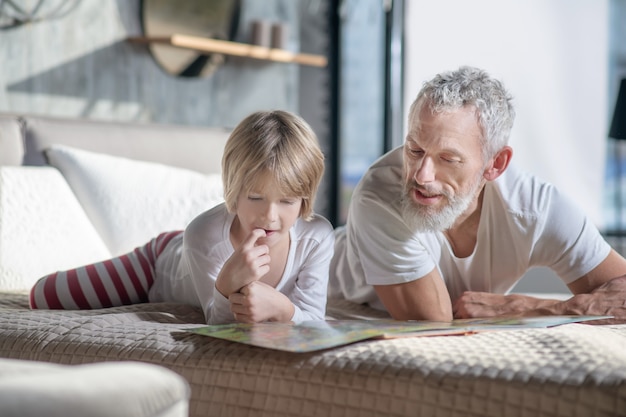 Loisirs, à la maison. Enfant attentif intéressé et papa barbu adulte allongé sur le lit en lisant à la recherche de livre