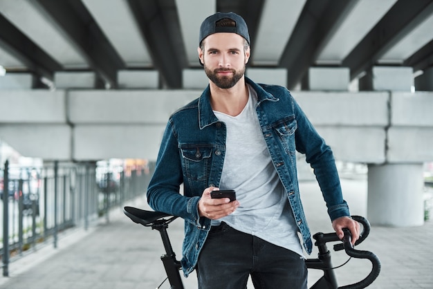 Loisirs de plein air jeune homme élégant assis sur un vélo dans la rue de la ville tenant un smartphone à la recherche