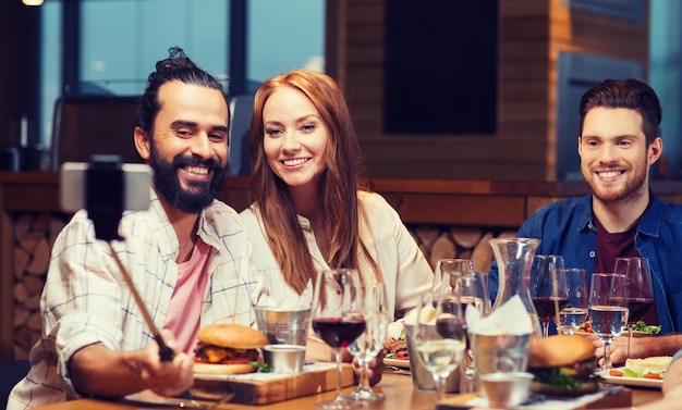 loisirs, technologie, amitié, gens et vacances concept - amis heureux dînant et prenant une photo par smartphone selfie stick au restaurant