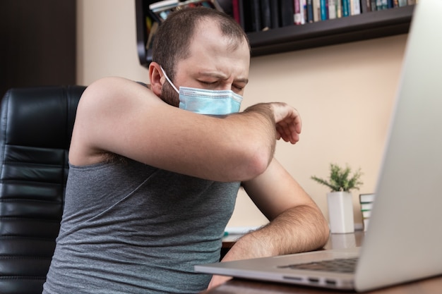 Loisirs utilisant un ordinateur portable. Un jeune homme barbu avec un masque dans les vêtements à la maison utilise un ordinateur portable