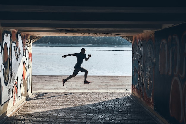 Long saut. Toute la longueur du jeune homme africain en vêtements de sport faisant du jogging tout en faisant de l'exercice près de la rivière à l'extérieur