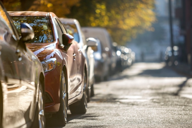 Longue rangée de différentes voitures et fourgonnettes brillantes garées le long de la route vide aux beaux jours d'automne sur fond de bokeh feuillage doré vert flou.