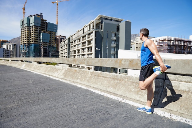 Longue route pour vaincre Jeune homme étirant ses jambes avant une course sur route dans la ville