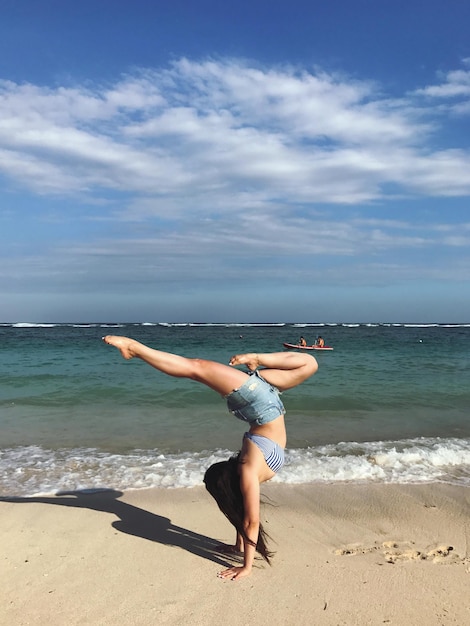 La longueur complète d'une femme faisant un handstand sur le rivage à la plage contre le ciel