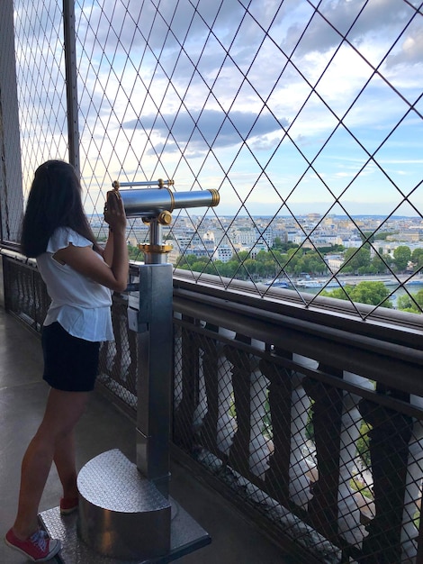 La longueur complète de la femme tenant des jumelles alors qu'elle se tient contre le ciel vu à travers la clôture