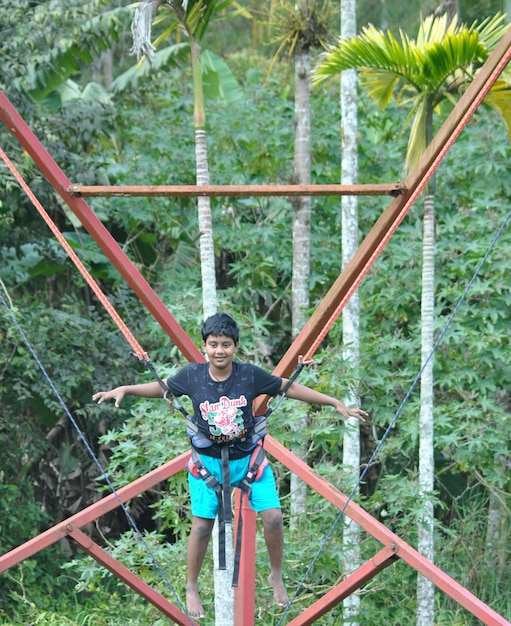 Photo longueur complète de garçon souriant suspendu avec un harnais de sécurité dans la forêt