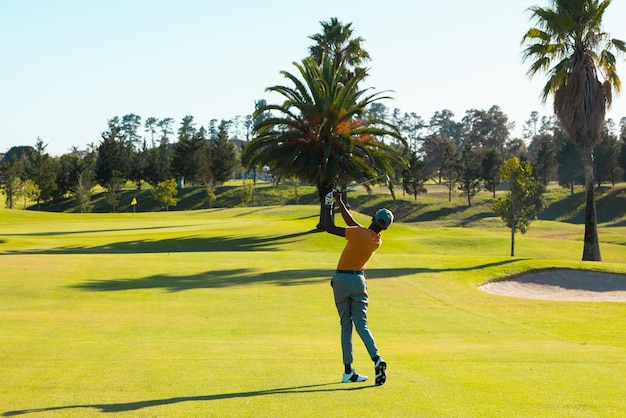 Photo longueur complète d'un jeune homme afro-américain jouant au golf contre des arbres et un ciel clair sur un terrain de golf