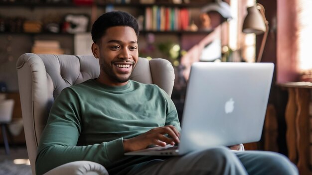 Photo une longueur complète de jeune homme noir souriant avec un ordinateur portable assis dans un fauteuil travaillant en ligne