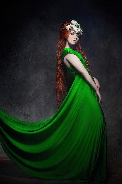 Look fabuleux de fille rousse, robe longue verte, maquillage lumineux et gros cils. Femme fée mystérieuse aux cheveux rouges. De grands yeux et des ombres colorées, de longs cils. Look sexy, princesse sur fond sombre