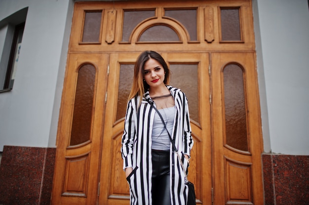 Look femme à la mode avec une veste de costume à rayures noires et blanches, un pantalon en cuir, posant contre la grande porte en bois du bâtiment. Concept de fille fashion.