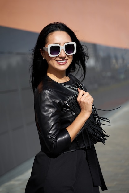 Look de mode de rue d'automne. Jolie femme dans des vêtements cool dans des lunettes de soleil élégantes. Modèle portant une robe noire et une veste en cuir à franges noires.