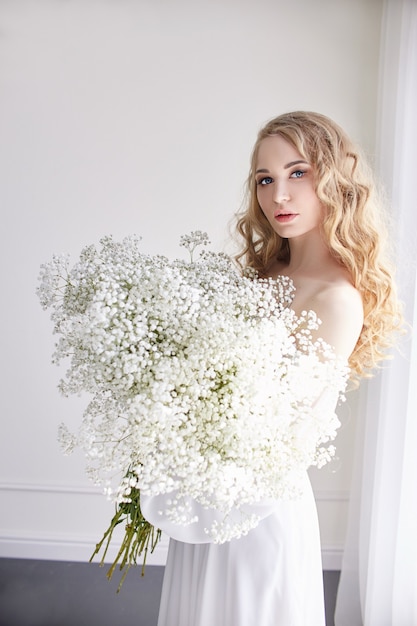 Look romantique blonde bouclée, beaux yeux. Fleurs sauvages blanches dans les mains. Fille robe légère blanche et cheveux bouclés