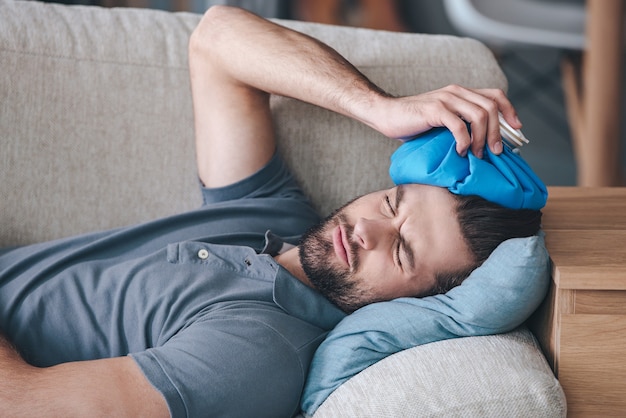 Lorsque vous frappez l'étagère avec votre tête. Jeune homme frustré tenant un sac de glace sur la tête en position allongée sur le canapé à la maison