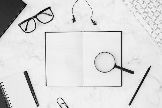 Photo des loupes sur le cahier entourées d'un clavier; lunettes; écouteur; crayon; et agenda sur fond texturé