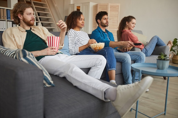 Low angle view at multi-ethnique groupe d'amis à regarder la télévision ensemble tout en étant assis sur un canapé confortable à la maison et en appréciant des collations