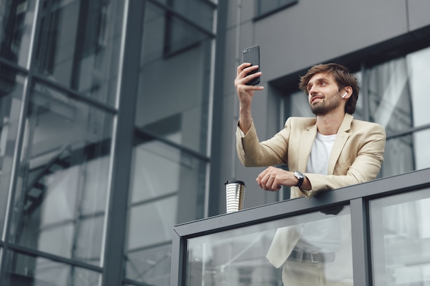 Low angle view of bel homme en tenue de soirée ayant un chat vidéo sur smartphone en se tenant debout sur le balcon du centre de bureau