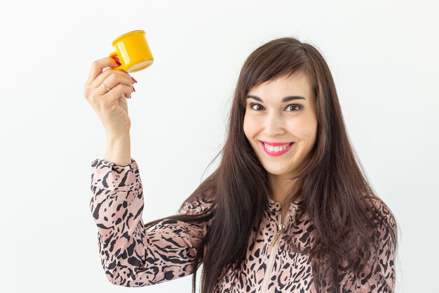 Ludique jeune femme brune tenant dans ses mains une petite tasse jaune posant sur un mur blanc