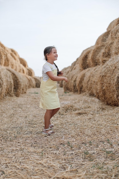 Ludique petite fille tressée brillante courant sur la route de campagne de foin entre les meules de foin regardant loin en robe d'été S'amuser loin de la ville sur un champ plein de foin doré