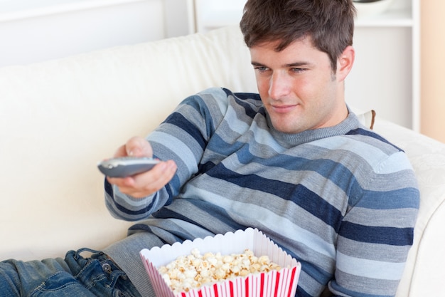 Photo lumineux jeune homme mangeant des pop-corn et tenant une télécommande couché sur