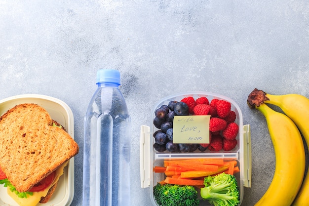 Lunchbox avec des baies de sandwich carottes brocoli bouteille d'eau banane sur fond gris