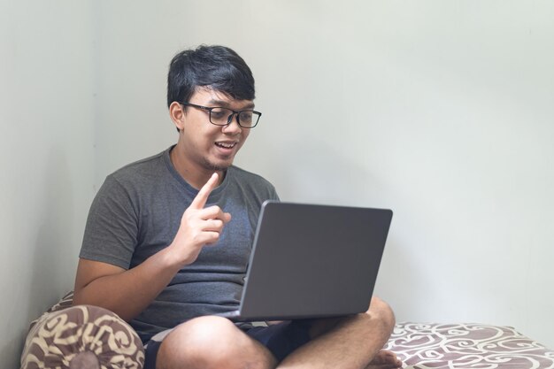 Photo lunettes asiatiques homme pointe l'index et regarde un ordinateur portable sur une chambre pour le travail à domicile concept avec lumière naturelle