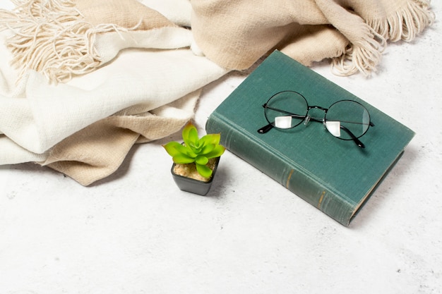 Des lunettes à lunettes rondes reposent sur le Livre, une écharpe en laine et une fleur de chambre sur fond blanc clair.