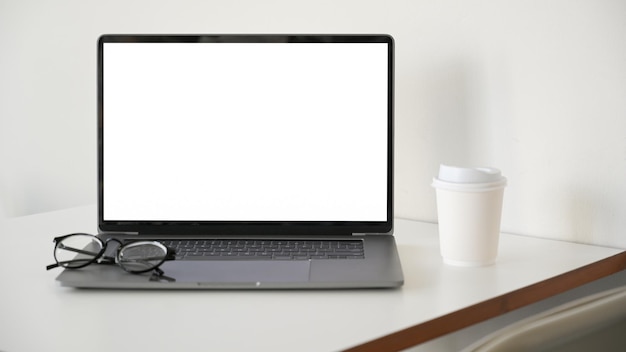 Des lunettes de maquette d'écran blanc d'ordinateur portable d'ordinateur portable et une tasse de café sont sur le bureau blanc