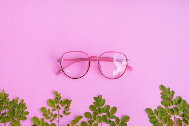 Lunettes roses transparentes sur fond de papier rose pastel avec accessoires de feuilles vertes