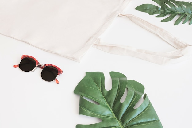 Lunettes de soleil, sac en coton et monstera vert sur fond blanc