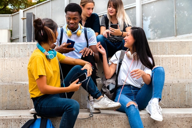 Des lycéens adolescents multiraciaux heureux sont assis sur les marches à parler et à utiliser des téléphones portables à l'extérieur.