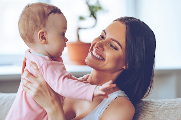 Ma petite fille! Joyeuse belle jeune femme tenant une petite fille dans ses mains et la regardant avec amour alors qu'elle était assise sur le canapé à la maison